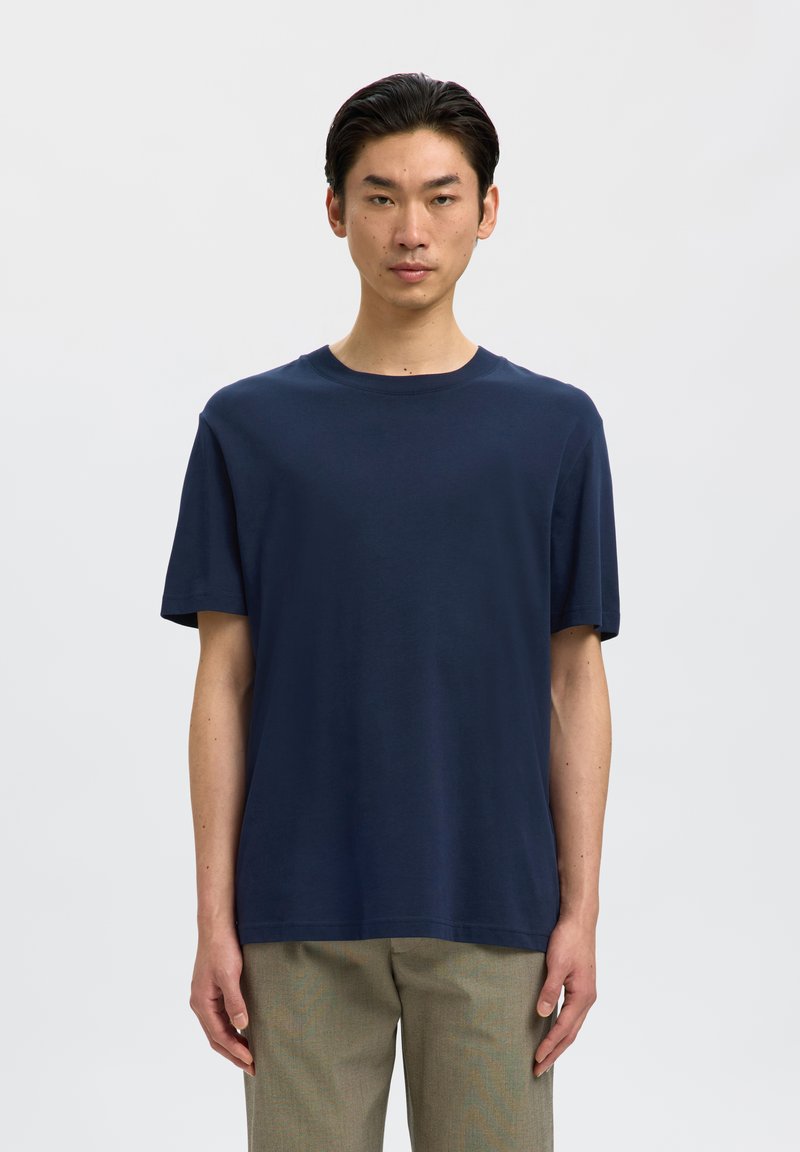 Joven con cabello corto y oscuro que lleva una camiseta lisa de manga corta azul marino y pantalones beige, de pie sobre un fondo blanco liso.