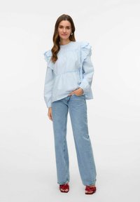 Blusa azzurra chiara con spalline a ruches, vita elasticizzata e maniche lunghe, abbinata a jeans azzurri chiari e sandali rossi.