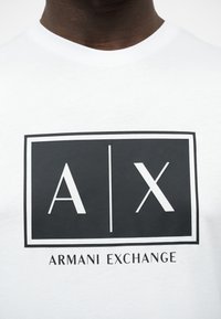 Κοντινό σε λευκό πουκάμισο με μαύρο ορθογώνιο λογότυπο που φέρει τα γράμματα "A|X" και το κείμενο "ARMANI EXCHANGE" από κάτω.