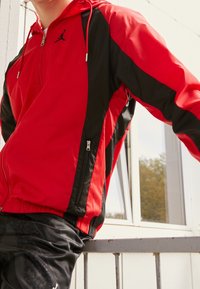Personne portant une veste à fermeture éclair rouge et noire avec une capuche et un pantalon de sport noir, appuyée contre une rambarde métallique près d'une fenêtre.