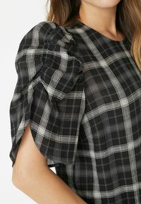 Top în carouri alb-negru, cu mâneci scurte bufante, țesătură texturată și un design lejer, evidențiind modelul în grilă și detaliile accent.