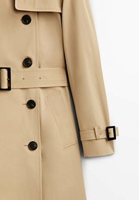 Trench-coat beige avec boutons noirs, taille ceinturée, poche latérale, manches longues et détail de sangle avec boucle au poignet.