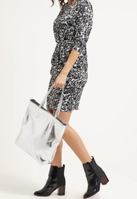 Sölvfärgad väska med två handtag, slät textur, och en mönstrad klänning i svart och vitt med trekvartsärmar, bärs med svarta ankelstövlar.
