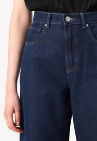 Σκούρο μπλε τζιν παντελόνι με ψηλή μέση, μπροστινές τσέπες και αντίθετη ραφή. Διαθέτει κλείσιμο με κουμπί και φερμουάρ.