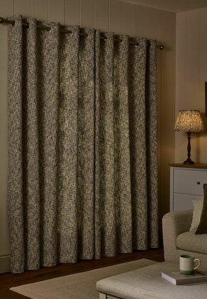 Thick textured beige curtains hanging from a metal rod beside a lit table lamp, beige sofa, and coffee table with a mug and book.