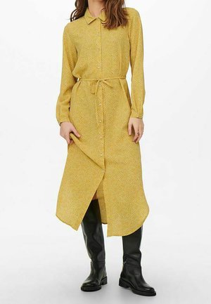 Robe chemise jaune moutarde à manches longues, taille cintrée et motif floral blanc, associée à des bottes noires hautes jusqu'aux genoux.