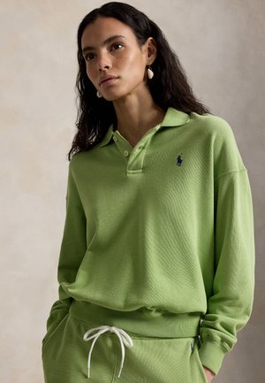 Femeie cu păr lung și închis la culoare, purtând un pulover polo verde deschis și pantaloni asortați cu șnur alb, stând în fața unui fundal simplu.