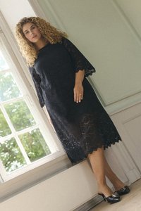 Robe en dentelle noire avec des manches cloche trois-quarts, un ourlet festonné et un motif floral. Associée à des talons noirs brillants avec un accent en forme de nœud.