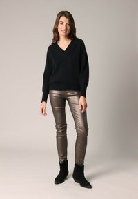 Jeune femme aux cheveux bruns portant un pull noir en V, un pantalon serré taupe brillant et des bottines noires, se tenant sur un fond uni.