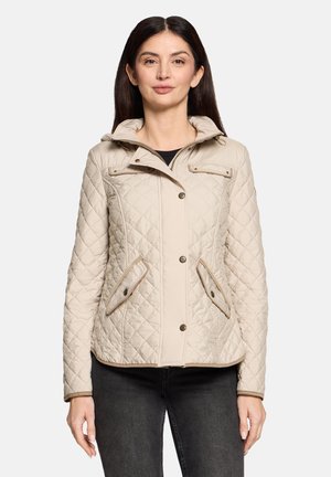 Femme portant une veste matelassée beige avec poches avant et boutons-pression, associée à un pantalon noir, debout devant un fond uni.