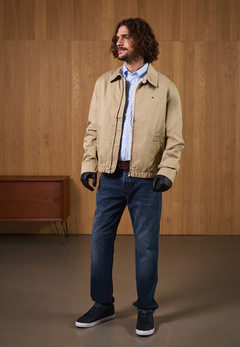 Beige bomber jacket over a blue plaid shirt, paired with dark jeans and black sneakers. Model wears black gloves and stands against wood backdrop.