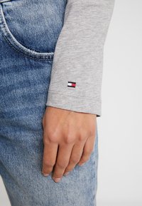 Szara koszulka z długim rękawem z prążkowanymi mankietami, z małym haftowanym logo w czerwonym, białym i niebieskim kolorze, w połączeniu z niebieskimi dżinsami.