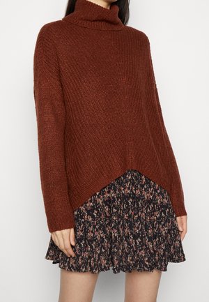 Femme portant un pull à col roulé marron ample et une jupe plissée noire à fleurs, debout devant un fond uni.