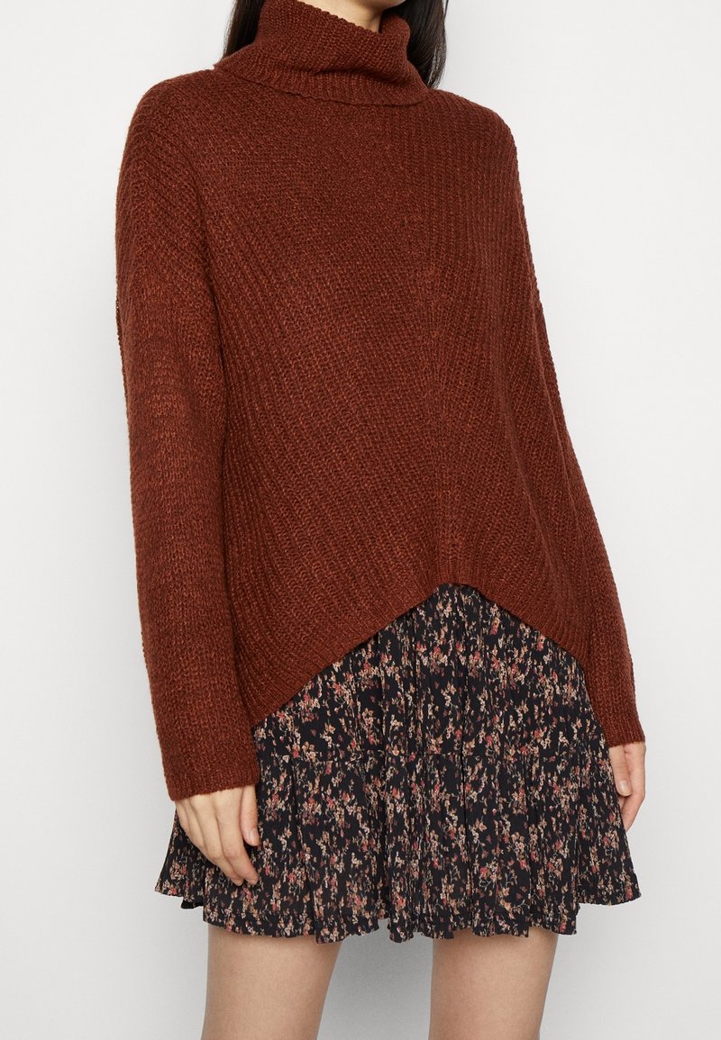 Femme portant un pull à col roulé marron ample et une jupe plissée noire à fleurs, debout devant un fond uni.