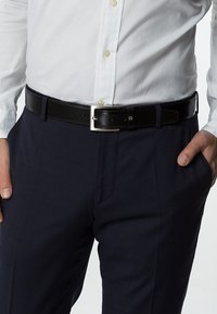 Cinturón de cuero negro con una hebilla rectangular plateada, usado sobre pantalones texturizados en navy, combinado con una camisa blanca de botones.