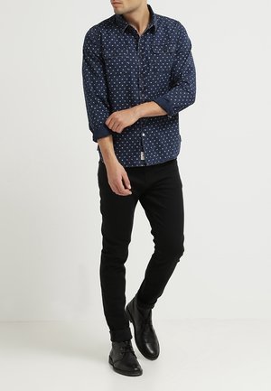 Uomo che sistema la manica di una camicia blu navy a motivi con bottoni, indossa pantaloni neri e scarpe nere, in piedi su sfondo bianco.