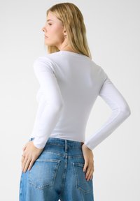 Uživeni bijeli top s dugim rukavima i okruglim izrezom u kombinaciji sa svijetloplavim trapericama s bočnim džepovima na stražnjoj strani i blagim oštećenjima.