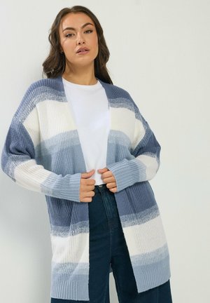 Femme portant un cardigan tricoté rayé bleu, blanc et gris sur une chemise blanche et un jean bleu foncé sur un fond uni.