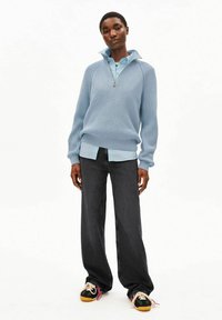 Lichtblauwe geribbelde pullover met een ritskraag, gedragen over een lichtblauw shirt, gecombineerd met wijde donkere jeans en sneakers in meerdere kleuren.