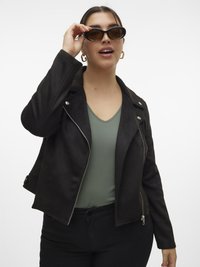 Femme ajustant des lunettes de soleil ovales noires, portant une veste zippée noire sur un t-shirt vert à col en V et un pantalon noir, avec des boucles d'oreilles créoles dorées.