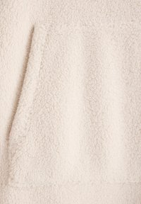 Fleece w kolorze kremowym o miękkiej, teksturowanej powierzchni. Materiał posiada wyraźną kieszeń i ma gruby, przytulny wygląd.