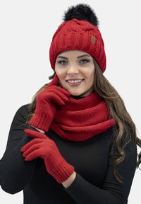 Rød strikket hue med en sort pom-pom, matchende rød tørklæde med rillede tekstur og røde handsker med rillede manchetter, der har en glat finish.