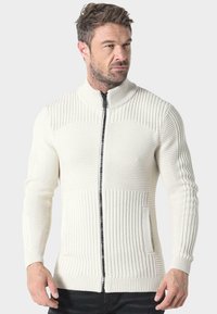 Suéter de punto color crema con un cuello alto, textura acanalada y cremallera frontal. Presenta un bolsillo lateral y mangas largas.