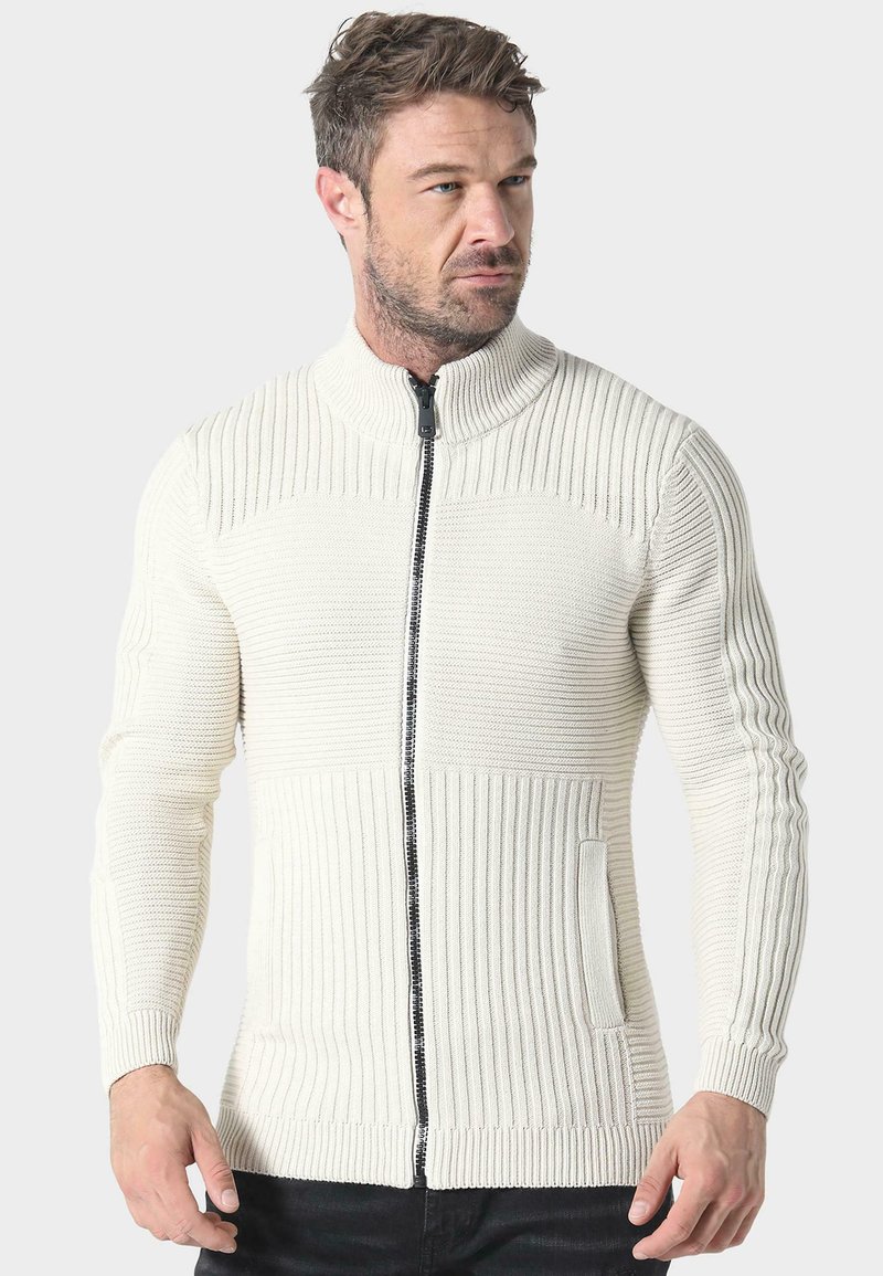 Suéter de punto color crema con un cuello alto, textura acanalada y cremallera frontal. Presenta un bolsillo lateral y mangas largas.