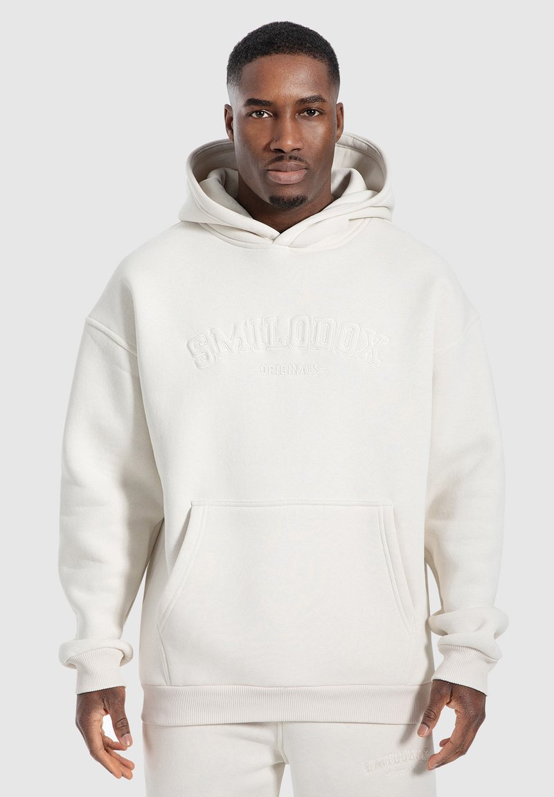 Ljusbeige hoodie med en känguruficka, oversized design, ribbade ärmslut och broderad logotyp på bröstet. Tillverkad av strukturerad tyg.
