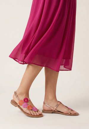 Nogi u prześwitującej magentowej spódnicy midi i płaskich sandałach w kolorze różowego złota, ozdobionych różowymi i pomarańczowymi aplikacjami w kształcie kwiatów.