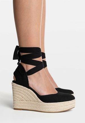Jambes portant des espadrilles compensées noires avec des liens à nouer autour de la cheville et des semelles plateformes en corde naturelle tressée sur fond blanc.