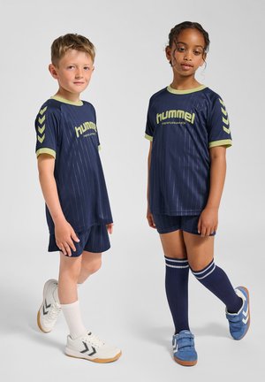 Marineblå sportsdragter med lodrette striber og limegrønne detaljer, kombineret med matchende shorts. Sneakers er hvide og blå med gummisåler.