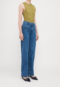 Top sin mangas texturizado en verde oliva con escote con volantes, combinado con jeans de mezclilla azul sueltos y de cintura alta y tacones negros puntiagudos.