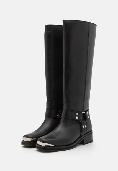 Botas negras de cuero hasta la rodilla con una textura suave, acentos plateados en la puntera, correa ajustable con tachuelas metálicas y un resistente tacón cuadrado.