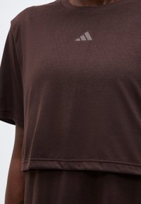 Σκούρο καφέ cropped μπλουζάκι με στρώσεις και μικρό αντανακλαστικό λογότυπο Adidas στο στήθος, φορεμένο από ένα άτομο.