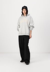 Grå hætte sweatshirt med et stort præget logo i lysegul, parret med sorte bredbenede bukser og en sort kasket med hvid tekst.