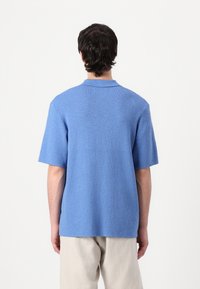 Polo din tricot albastru cu mânecă scurtă, având un finisaj texturat, cu guler plat și o croială lejeră, asortat cu pantaloni bej deschis.