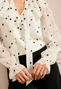 White blouse featuring black heart patterns, ruffled neckline, tie closure, and gathered sleeves. Smooth, sheer fabric with a soft texture.