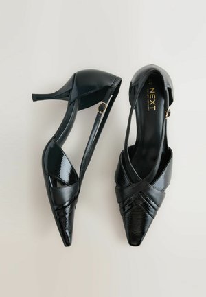 Fekete lakk magassarkú cipő, keresztpántos rögzítéssel, hegyes orral és oldalsó csattal. Simává bontott textúrával és fényes felülettel.