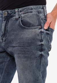 Un jean en denim bleu délavé présente une coupe structurée avec une poche visible et une fermeture à bouton en métal. Tissu texturé avec une légère élasticité.