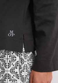 Camisa negra de manga larga con dobladillo y pequeña abertura, y texto blanco "M O'P", usada sobre pantalones con estampado floral en blanco y negro.