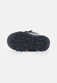 Suela de goma negra con patrón de textura y orificios de aire para transpirabilidad. Presenta una caja de dedos más ancha y acentos laterales reforzados.