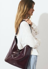 Burgundy leather shoulder bag with stud accents, slouchy design, and V-shaped top, featuring a smooth texture and adjustable strap.