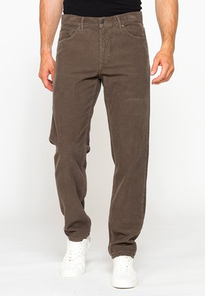 Uomo con pantaloni di velluto a coste marroni e scarpe da ginnastica bianche in piedi davanti a uno sfondo bianco semplice.