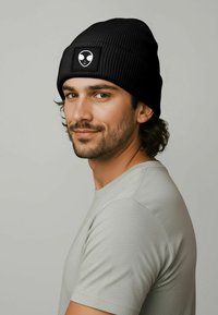 Black ribbed beanie with a square patch featuring a white alien graphic. Worn by a man in a light gray, textured shirt. Short, dark hair.