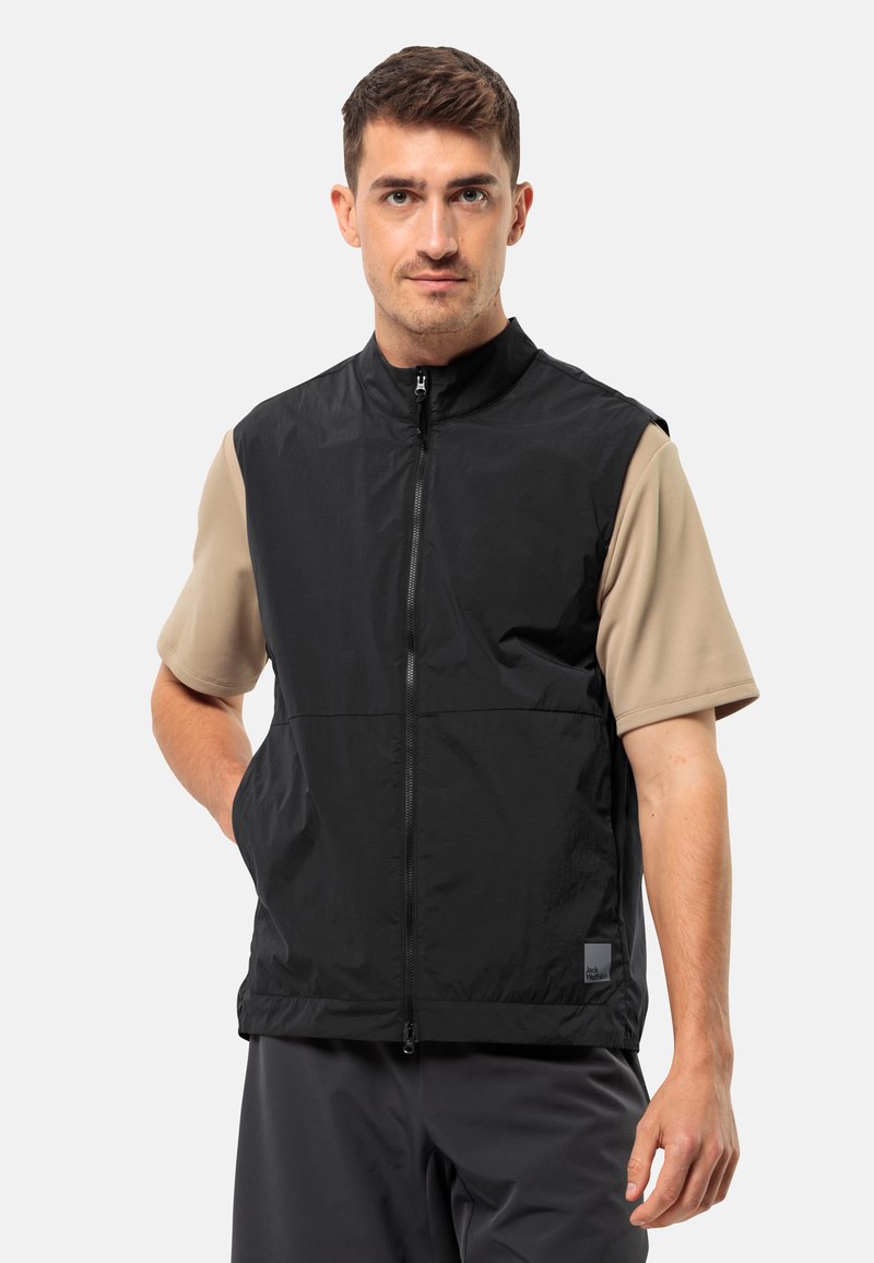Gilet nero in nylon con colletto alto e zip completa, dotato di tasche frontali e un'etichetta sul lato sinistro in basso. Indossato sopra una camicia beige.