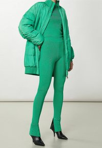 Groene pufferjack met hoge kraag, gecombineerd met een bijpassende fitted top en leggings met textuurpatronen. Zwarte puntige hakken.