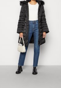 Černá péřová bunda s kapucí vystlanou kožešinou, bílé kostkované tričko, modré džíny rovného střihu a černé kotníkové boty, doplněné malou bílou kabelkou.