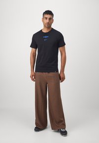 Black cotton t-shirt with blue logo, paired with wide brown plaid trousers. The outfit features a relaxed fit and soft texture.