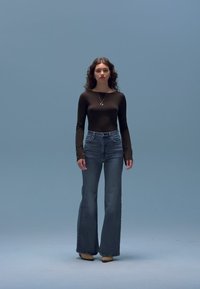 Top preto de manga comprida, jeans azul claro com corte evasê e saltos beges. Modelo está em frente a um fundo azul claro. Colar simples de prata.